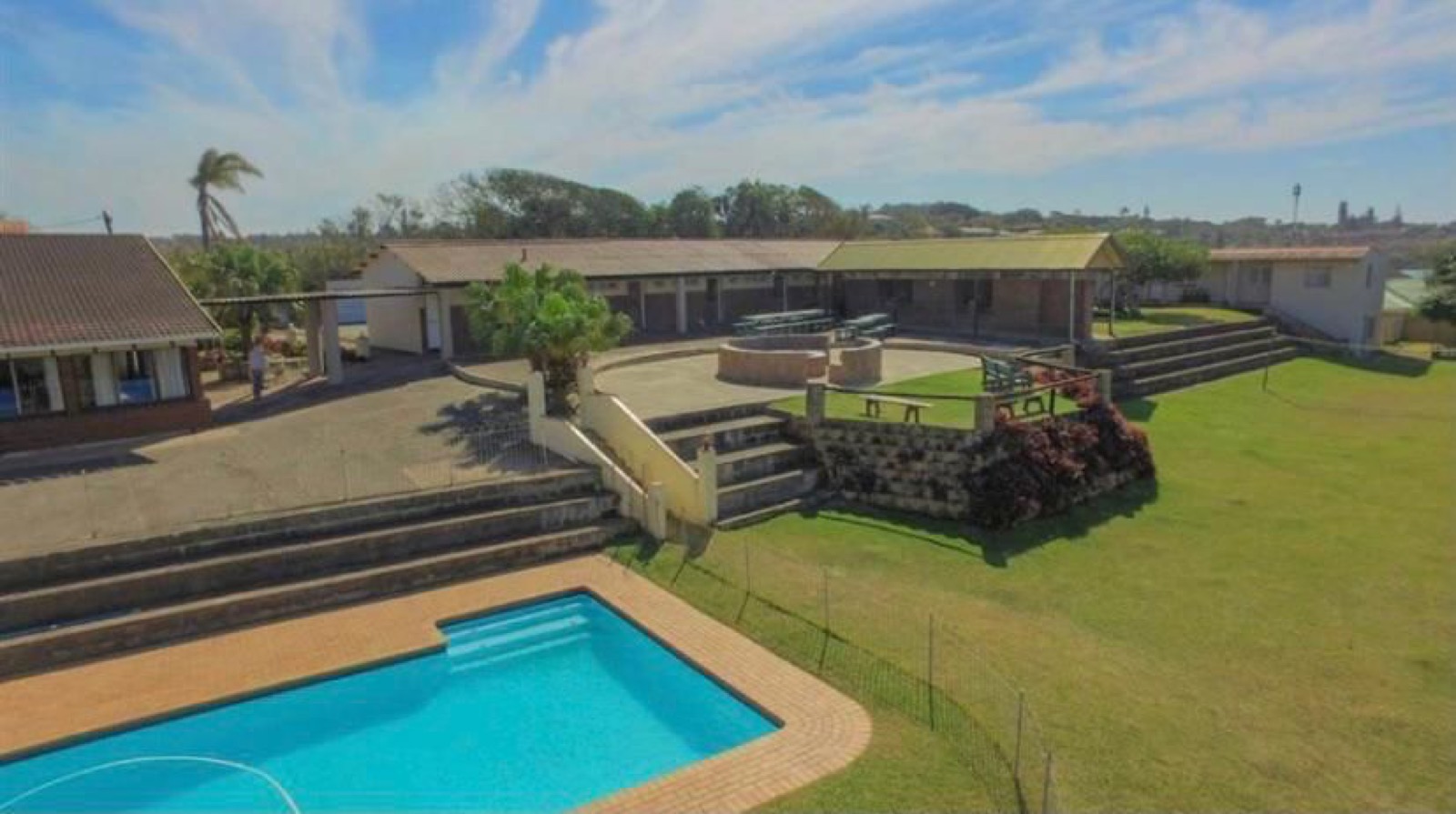 Aerial view of Tetelestai Recovery Centre showing swimming pool, manicured lawns, outdoor boma seating area, and open residential buildings under clear skies